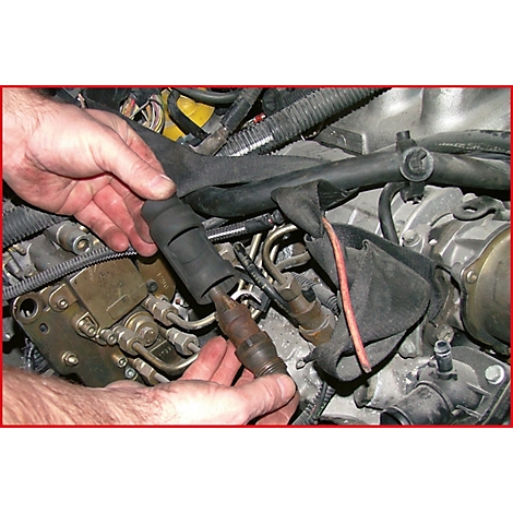 Nahaufnahme der Hände, die einen defekten Zündstecker aus einem Motor ausbauen. Sichtbar sind Schläuche, Kabel und weitere Motorteile.